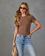 Woman wearing fitted brown short-sleeve crop top with light wash high-rise flare jeans, gold accessories, and brown sunglasses