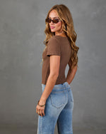 Woman modeling brown fitted crop tee and light wash high-waist jeans, wearing tortoise sunglasses, gold jewelry, gray background