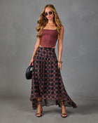 A woman models a two-piece outfit consisting of a sleeveless dusty rose crop top and a high-waisted midi skirt with a dark red and black geometric grid pattern. She is wearing black strappy heels, sunglasses, and holding a black woven handbag against a gray studio backdrop.
