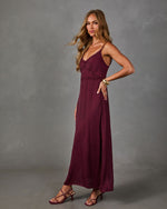 Woman in a burgundy satin maxi slip dress with lace trim and side slit, styled with strappy heeled sandals and gold bracelets