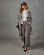A woman models a stylish outfit consisting of a brown and beige plaid coat over a white crop top, paired with wide-leg gray trousers, a brown cap, and sunglasses, set against a textured gray studio background.