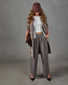A woman models a complete autumn outfit consisting of a brown plaid open-front coat over a white ribbed crop top, paired with gray wide-leg trousers, a brown cap, and dark brown pointed-toe heels, set against a neutral gray studio backdrop.