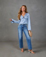 Woman wearing light blue satin tie-front long sleeve top with wide-leg medium wash jeans, clear heels, and woven clutch
