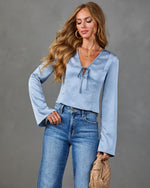 Woman wearing light blue satin V-neck top with bell sleeves and tie detail, paired with high-waist blue jeans and woven clutch