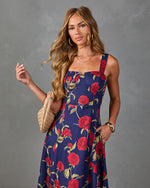 Navy floral maxi dress with red roses, square neckline, worn by woman with woven beige clutch and gold cuff bracelet