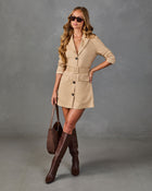 A woman models a beige belted blazer-style mini dress with dark buttons, styled with brown knee-high boots, a brown leather tote bag, and sunglasses against a textured gray studio backdrop.