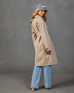 Woman wearing beige belted trench coat, wide-leg light wash jeans, gray Hamptons baseball cap, viewed from behind