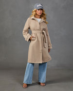Woman wearing belted tan trench coat, wide-leg jeans, gray Hamptons cap, and nude sandals on gray background