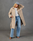 Woman wearing open beige trench coat with gingham lining, white ribbed top, wide-leg jeans, gray cap, and strappy sandals