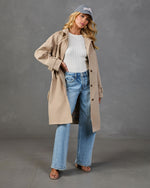 Woman wearing beige trench coat, white ribbed tank, wide-leg light wash jeans, strappy heels, and gray baseball cap