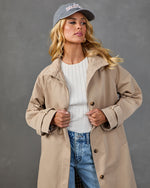 Woman modeling open tan trench coat over white ribbed top and blue jeans, wearing gray baseball cap, studio shot