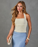 Woman in cream ruched halter top and blue satin midi skirt holding white clutch with gold hardware, layered gold jewelry