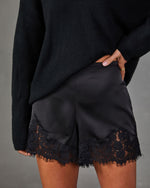 Black satin shorts with scalloped lace hem, styled with an oversized black knit sweater, hand resting on hip