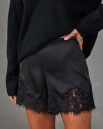Black satin high-waisted shorts with scalloped lace hem, styled with an oversized black knit sweater and gold rings