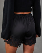 Back view of black satin shorts with elastic waist and scalloped lace hem, styled with a black cropped knit sweater