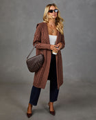 A woman models a long brown cable-knit cardigan with a hood, styled with a white top, dark blue jeans, brown heels, and a brown quilted shoulder bag against a gray studio backdrop.