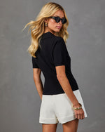 Woman modeling black ribbed knit polo top and white cuffed shorts with gold bangle bracelet and cat-eye sunglasses