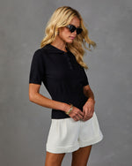Woman wearing black knit polo sweater top and white tailored cuffed shorts with gold bangle bracelet and cat-eye sunglasses
