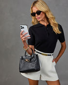 Woman in black knit polo top and white shorts holding phone, carrying black woven leather handbag with gold hardware