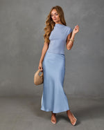 Woman wearing powder blue two-piece set: cap sleeve crop top and satin midi skirt, styled with woven mini bag and gold bangles