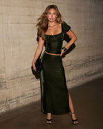 Woman in a green velvet puff-sleeve crop top and matching maxi skirt with side slit, styled with black heels and a fuzzy clutch