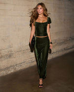 Woman in olive green velvet puff-sleeve crop top and matching maxi skirt with black heels and fur clutch against concrete wall