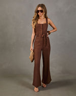 Brown linen wide-leg jumpsuit with square neckline, tie waist, and patch pockets styled with neutral sandals and woven clutch