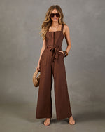 Brown wide-leg linen jumpsuit with square neckline, tie waist, and patch pockets, styled with nude sandals and woven bag
