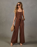 Woman wearing chocolate brown wide-leg linen jumpsuit with belt, square neckline, patch pockets, styled with nude heels and woven clutch