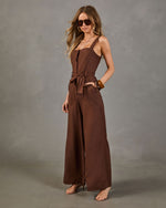 Brown wide-leg linen jumpsuit with corset bodice, tie waist, and square neckline styled with strappy heels and sunglasses