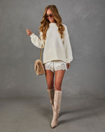 Woman in cream oversized knit sweater, satin lace-trim shorts, tan suede knee-high boots and faux fur crossbody bag