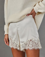 Cream satin shorts with scalloped floral lace hem, styled with an oversized ivory knit sweater against a gray backdrop