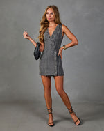 Woman wearing gray acid wash denim mini dress with deep V-neck, button front, raw hem, styled with black strappy heels and gold cuff bracelet.