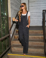 Woman in dark wash denim overalls and white tee holding iced coffee on outdoor stairs, black sunglasses, wavy blonde hair