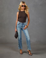 Woman wearing brown ribbed sleeveless bodysuit, light wash straight-leg jeans with frayed hem, black heeled sandals, holding dark woven clutch