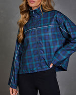 A woman wearing a cropped, long-sleeve windbreaker jacket with a blue and green plaid pattern and thin white piping details, paired with black pants against a gray studio background.