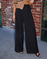 A person wearing high-waisted, wide-leg black pants with a beige knit cropped cardigan and black sandals, standing in front of a red brick wall.