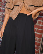 A close-up of a person wearing black wide-leg trousers and a cropped tan knit cardigan with black stripes, standing against a red brick wall.