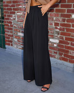 A person wearing high-waisted, wide-leg black pants with a tan textured crop top and black slide sandals, standing in front of a red brick wall.