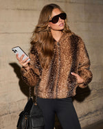 Woman in a brown spotted faux fur zip-up jacket, black sunglasses, gold hoops and black quilted bag, holding a phone