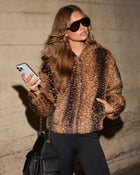 Woman in a brown spotted faux fur zip-up jacket, black sunglasses, gold hoops and black quilted bag, holding a phone