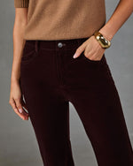 A close-up of a person wearing dark burgundy corduroy pants, styled with a tan sweater and a chunky gold cuff bracelet, against a neutral gray background.