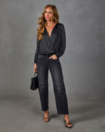 Woman in black satin wrap blouse, faded black barrel-leg jeans and strappy heels, holding a woven black handbag