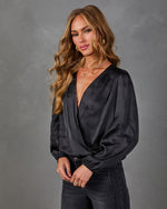 Woman in a black satin surplice blouse with balloon sleeves, layered gold necklaces and dark high-waisted jeans against gray backdrop