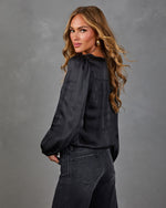 Woman looking over her shoulder in a black satin balloon-sleeve blouse and dark wide-leg jeans with gold hoop earrings