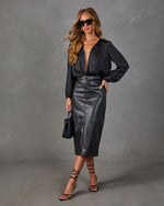 Woman in black satin plunge blouse, faux leather midi skirt, belt, strappy lace-up heels and woven handbag against gray backdrop