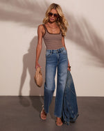 Woman wearing brown striped tank bodysuit, wide-leg jeans, sandals, holding woven bag and denim jacket, summer casual style
