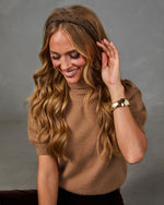 A woman with long wavy blonde hair wearing a brown polka-dot headband, a brown knit turtleneck sweater, and a gold cuff bracelet, posing against a neutral gray studio background.