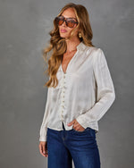 Woman wearing a cream satin button-front blouse with V-neck and long sleeves, styled with dark blue jeans and tinted aviators