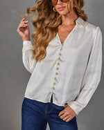 Woman wearing a cream satin long-sleeve button-front blouse with V-neckline and gold buttons, styled with dark denim jeans